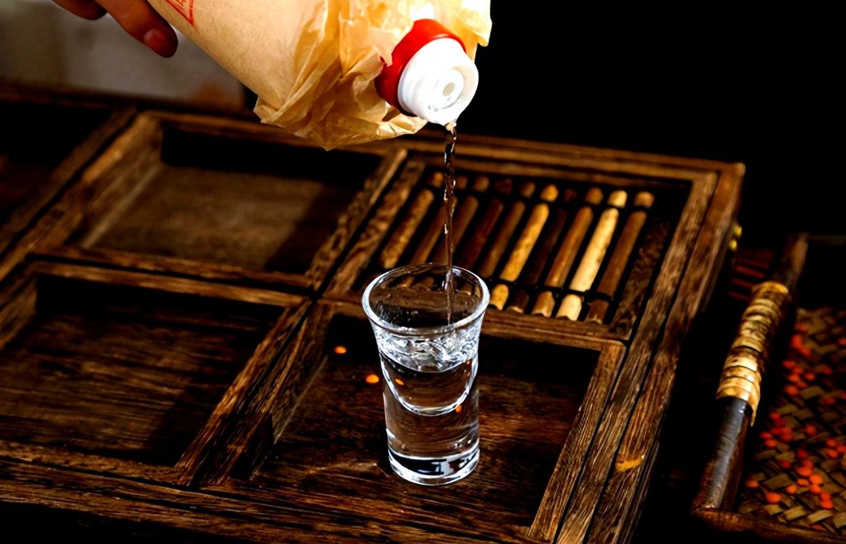 Clear baijiu pouring from a paper-wrapped bottle into a small heavy-based glass on a dark wooden tray