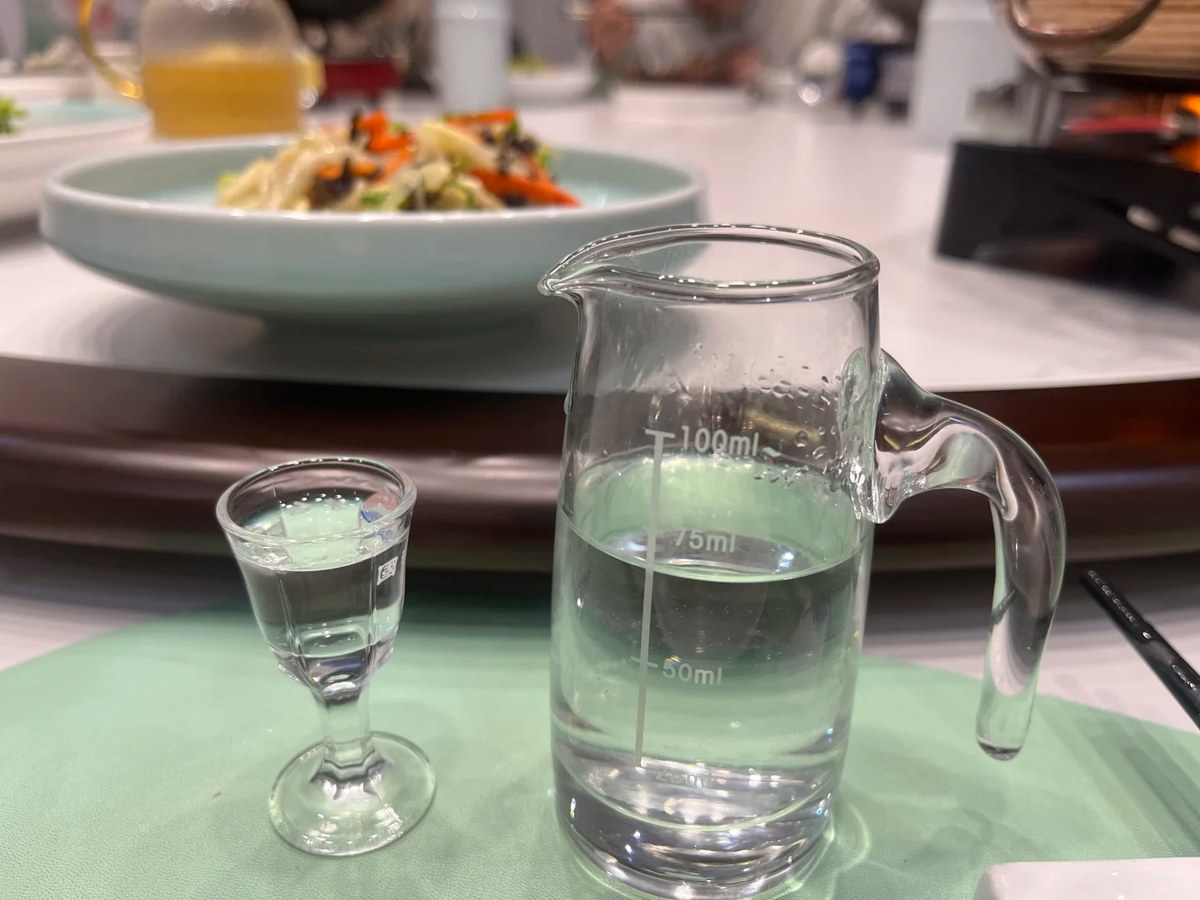 A baijiu tasting glass and a graduated pouring pitcher on a restaurant table, a bowl of 100ml marks away from the next toast