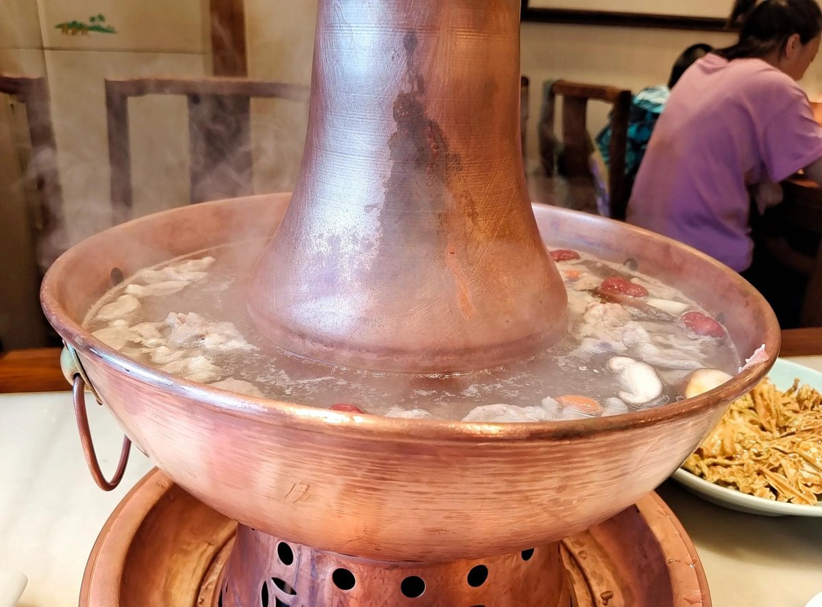 Beijing mutton hot pot — a copper chimney pot with charcoal glowing at its core, set on a restaurant table with broth simmering in the ring around it