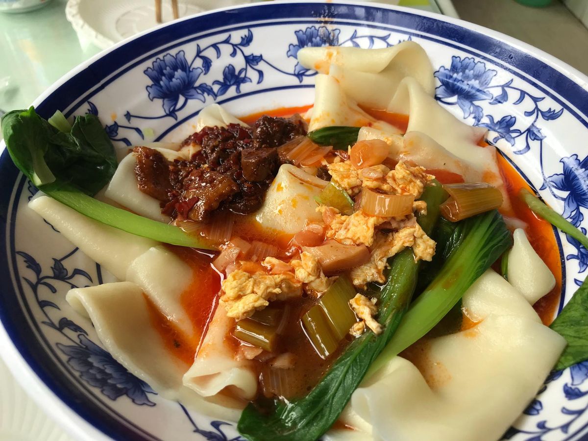 Biang Biang Noodles — a bowl of wide Shaanxi belt noodles in chili oil with greens and toppings, at a restaurant table in Xi'an