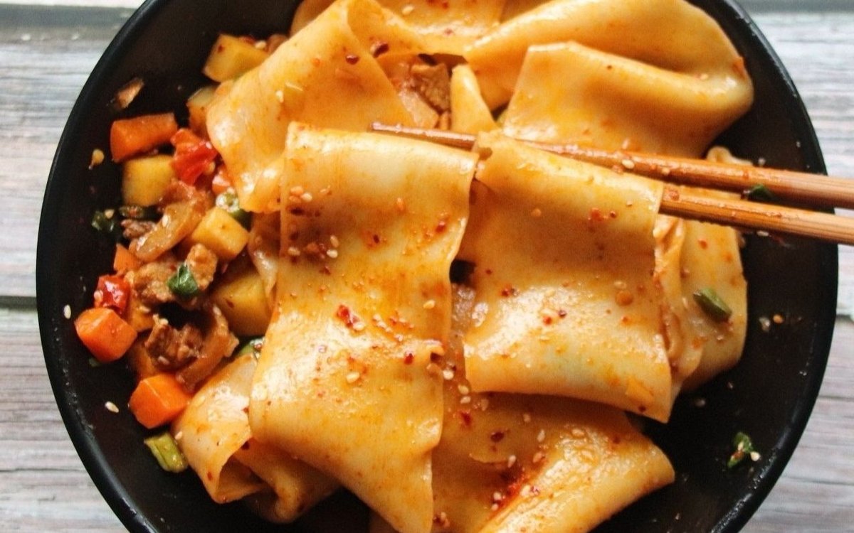 Biang Biang Noodles close-up — wide hand-pulled belt noodles coated in chili oil, sesame and chili flakes, lifted with chopsticks