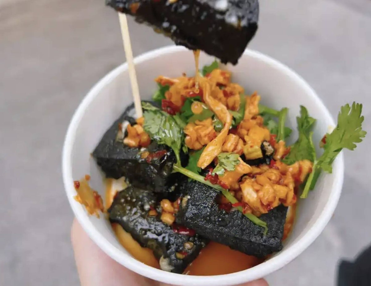 Changsha stinky tofu in a paper cup, one cube lifted out on a bamboo skewer