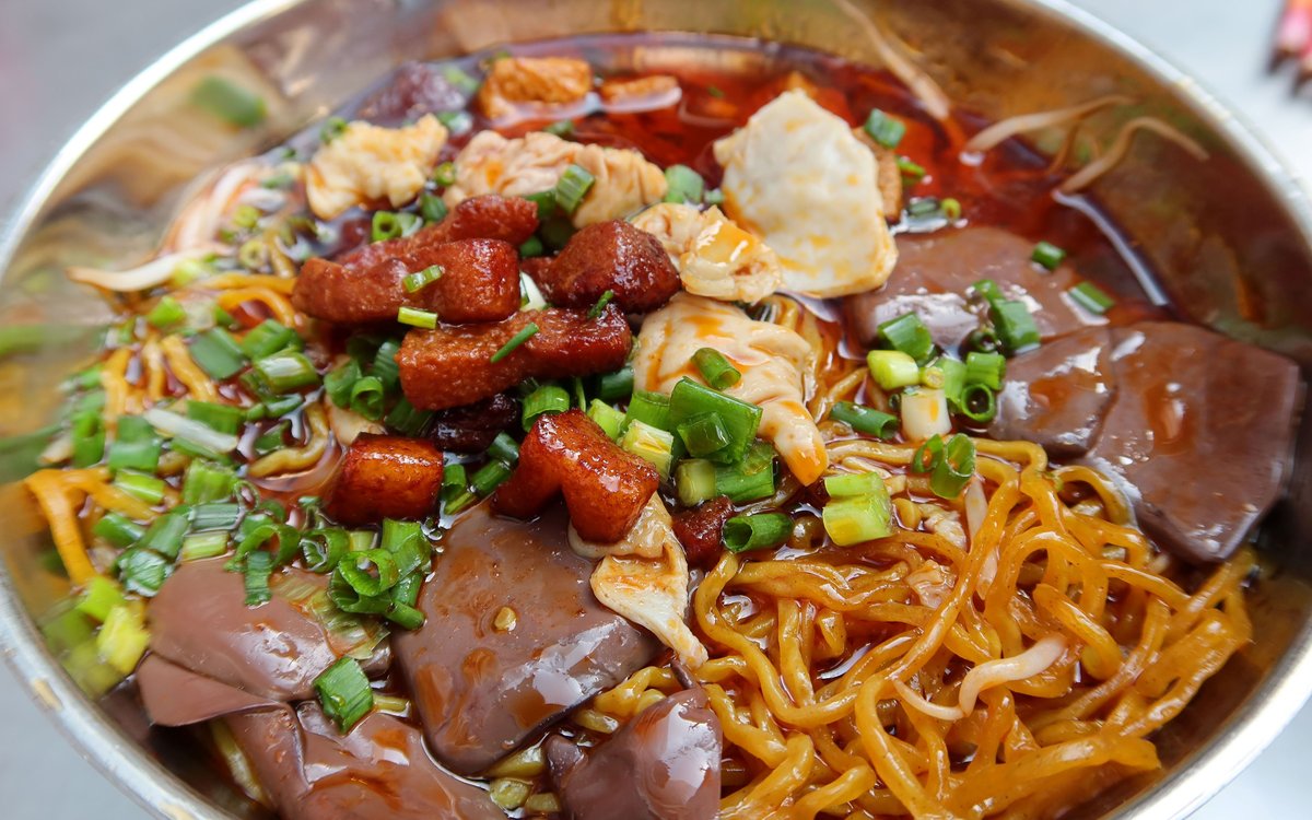 Close-up of a changwang mian bowl — thin yellow egg noodles, glossy cubes of congealed blood, braised pork intestine and chopped scallions in bright red chili oil