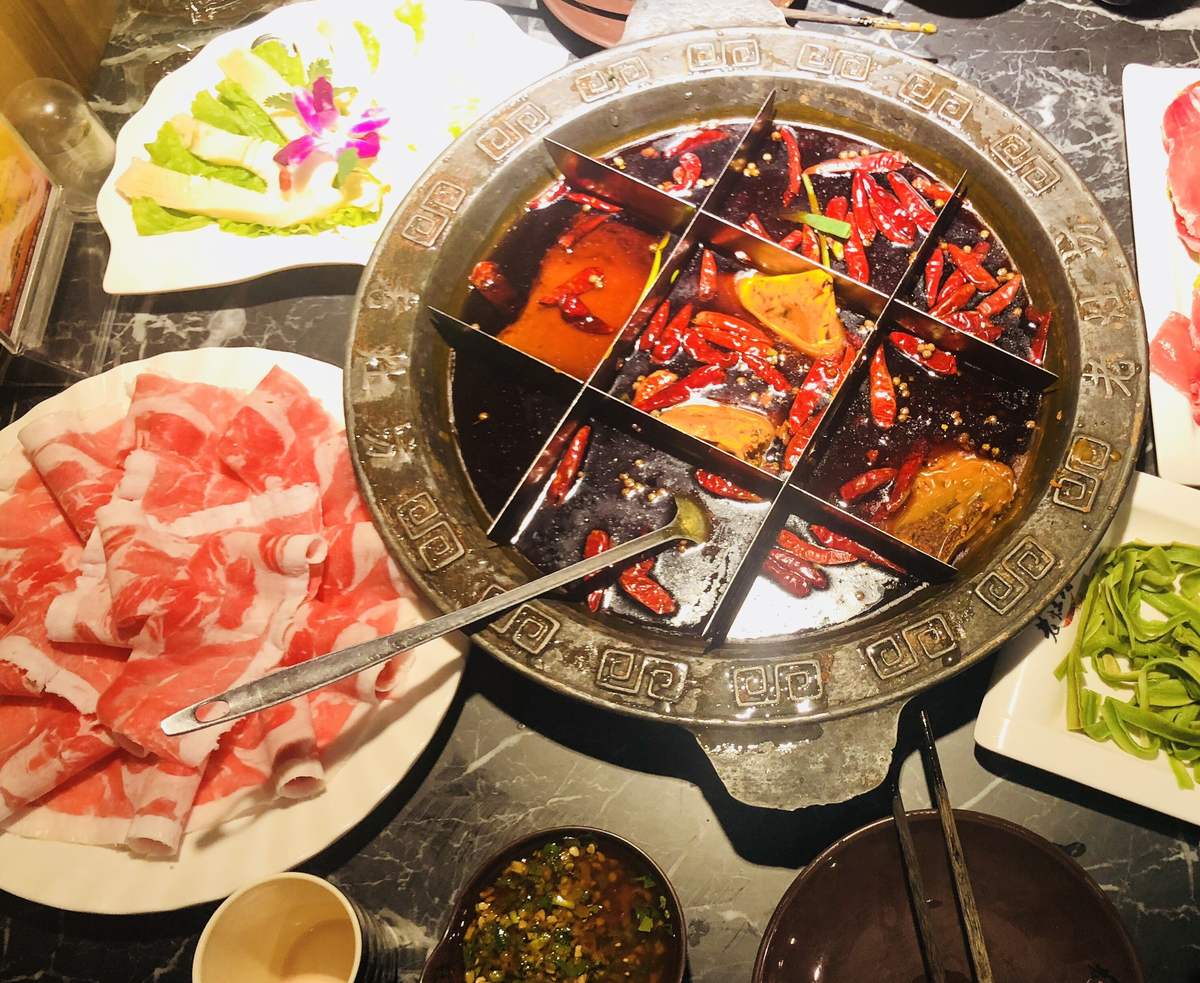 Chongqing hot pot — a nine-grid iron cauldron of dark beef tallow broth crusted with dried chilies, ringed by plates of raw beef and greens