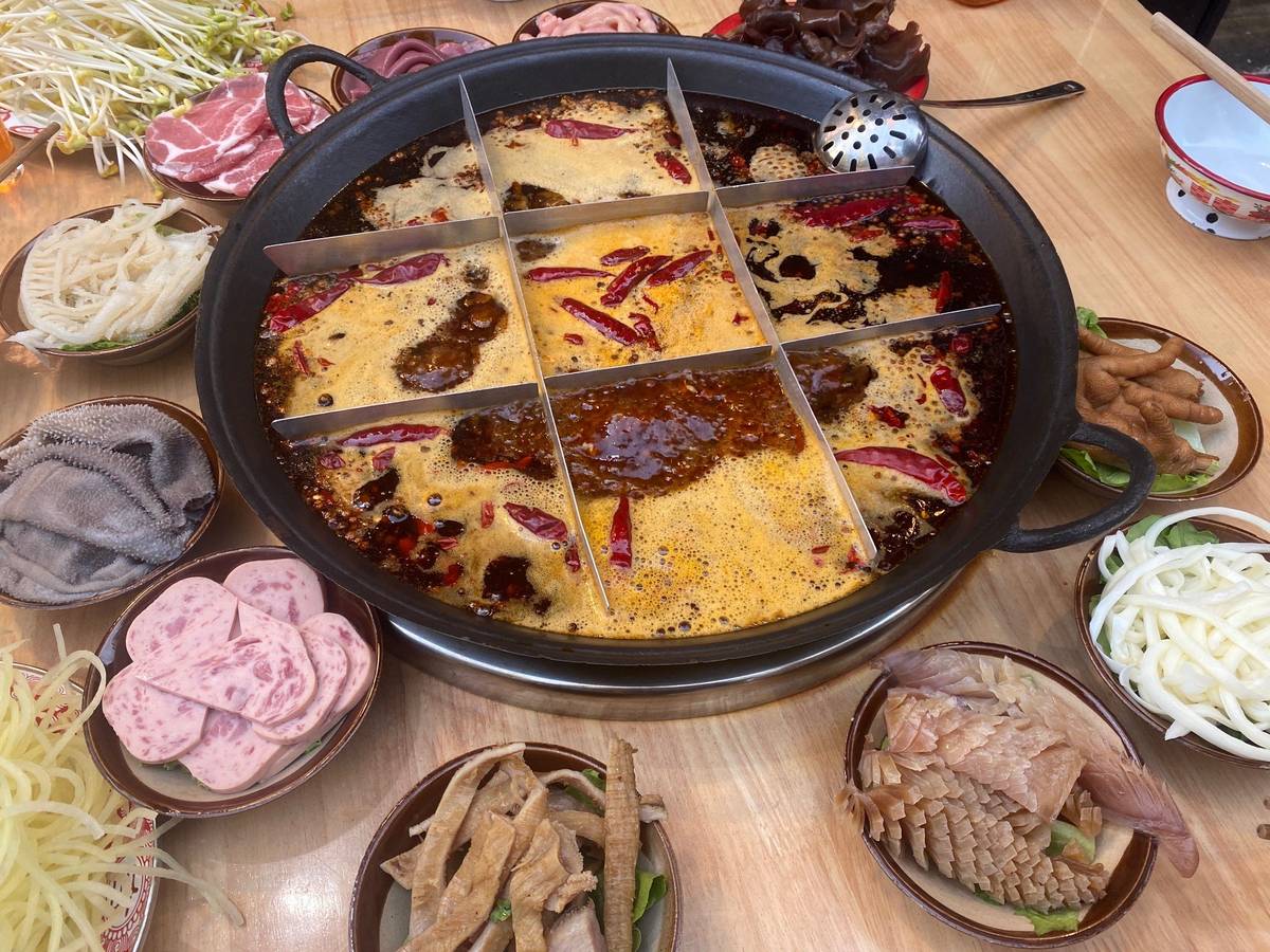 Red beef tallow broth at a full boil — dried chilies and chili sediment breaking on the surface, plates of tripe and luncheon meat crowding the table