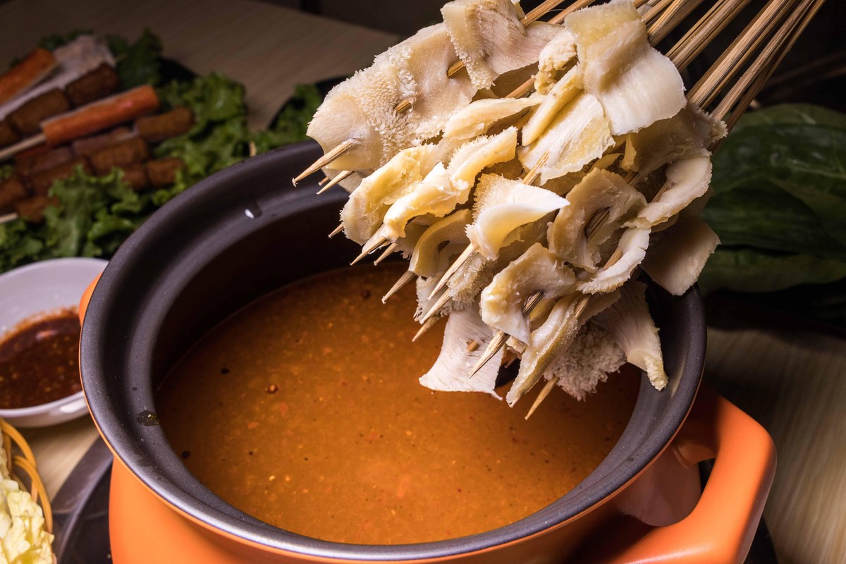 Skewered honeycomb tripe lowered into the boiling broth — pale frills about to curl in the red oil