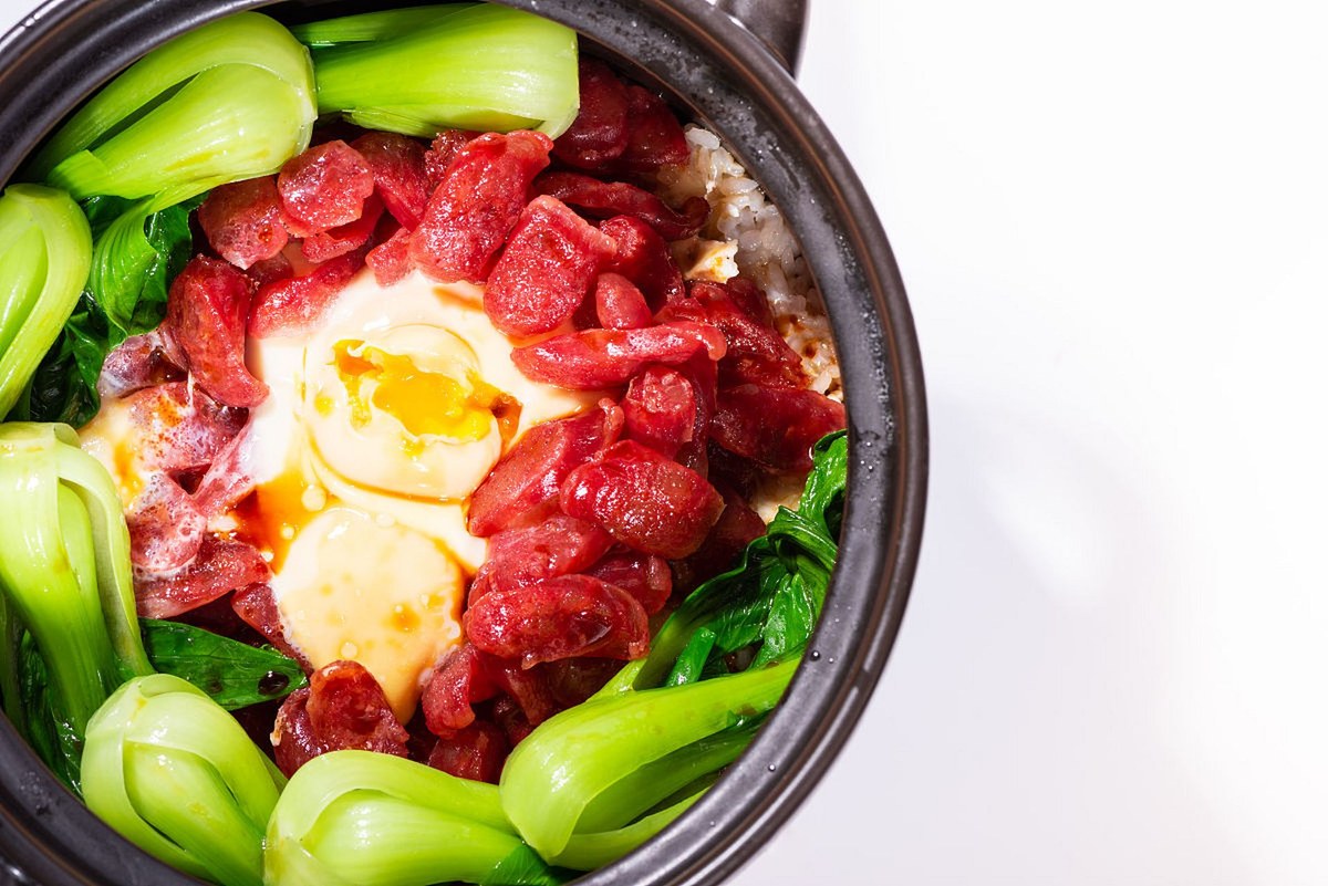 Cantonese Claypot Rice — a whole pot of rice under cured sausage, egg and greens, the lid just off
