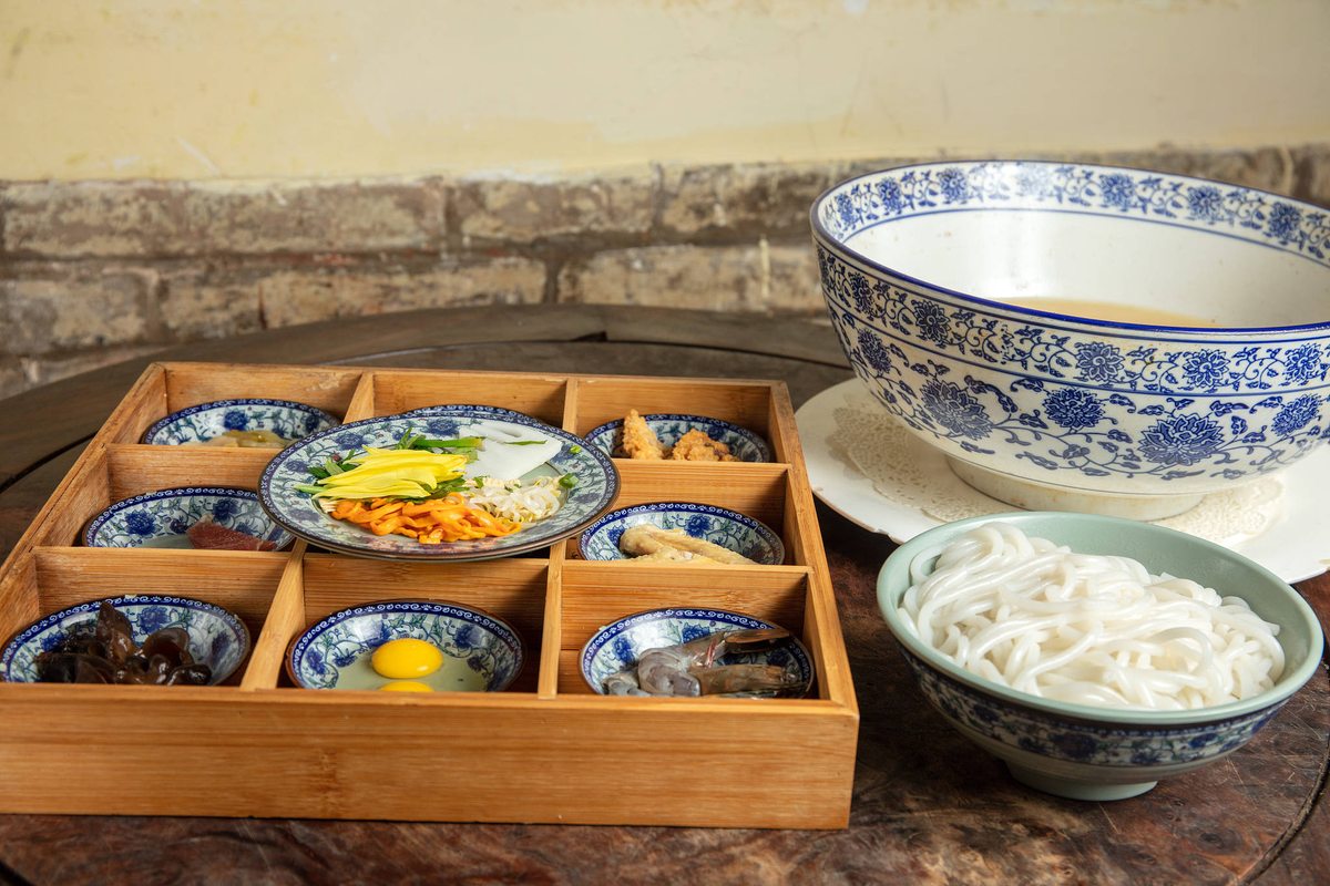 Crossing-Bridge Noodles — a Yunnan set on dark wood: a bowl of still broth, a nest of rice noodles, and a wooden tray of small dishes of raw meat, egg and greens