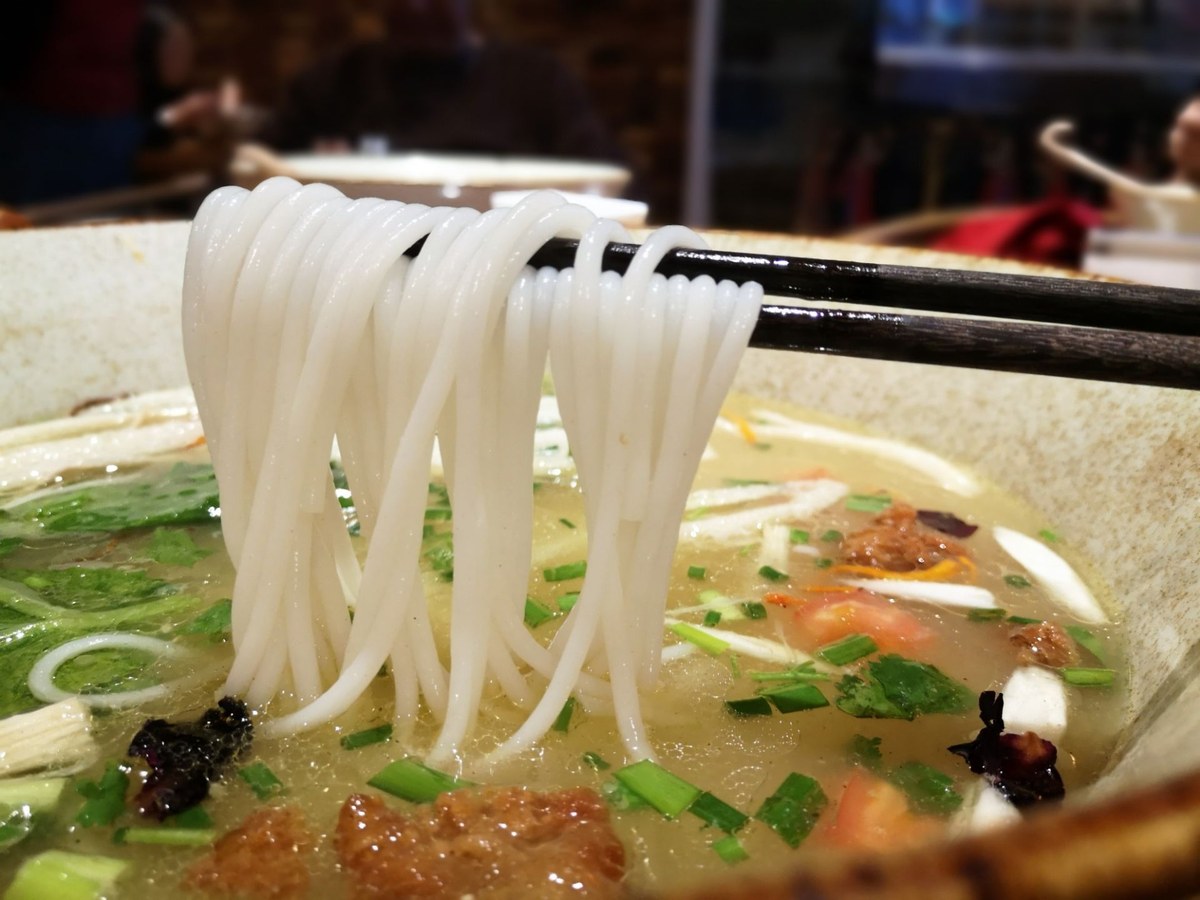 Crossing-Bridge Noodles close-up — chopsticks lifting a bundle of thick white rice noodles out of the broth