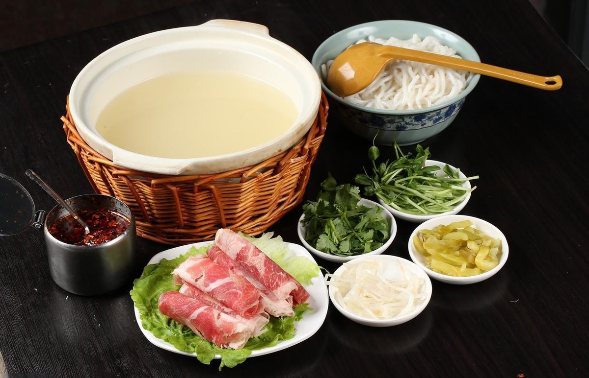 Crossing-Bridge Noodles served at the table — a pot of milky broth in a woven basket, a bowl of rice noodles, and plates of raw meat and garnishes