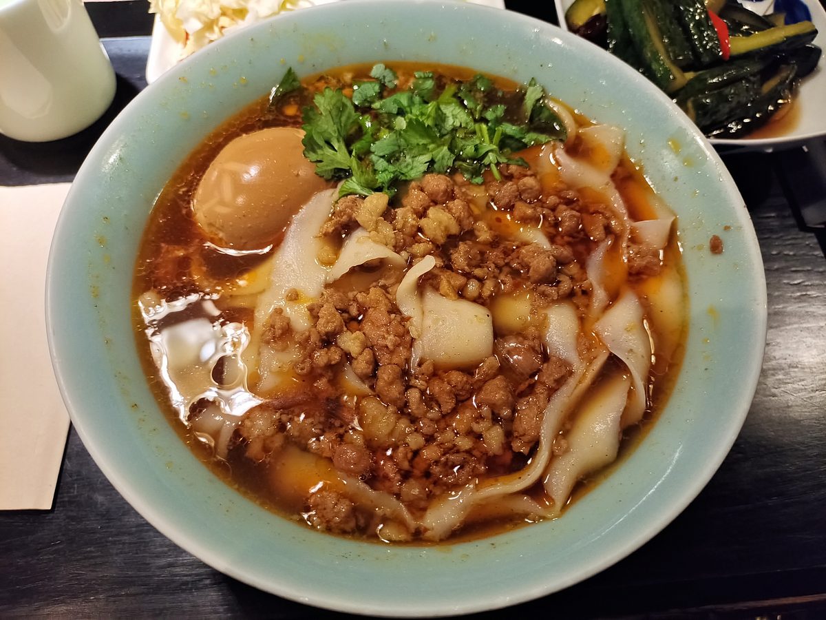 Shanxi sliced noodles — a pale blue bowl of thick irregular noodles under a dark braise with minced meat, a braised egg and coriander, side dishes pushed to the edge of the table