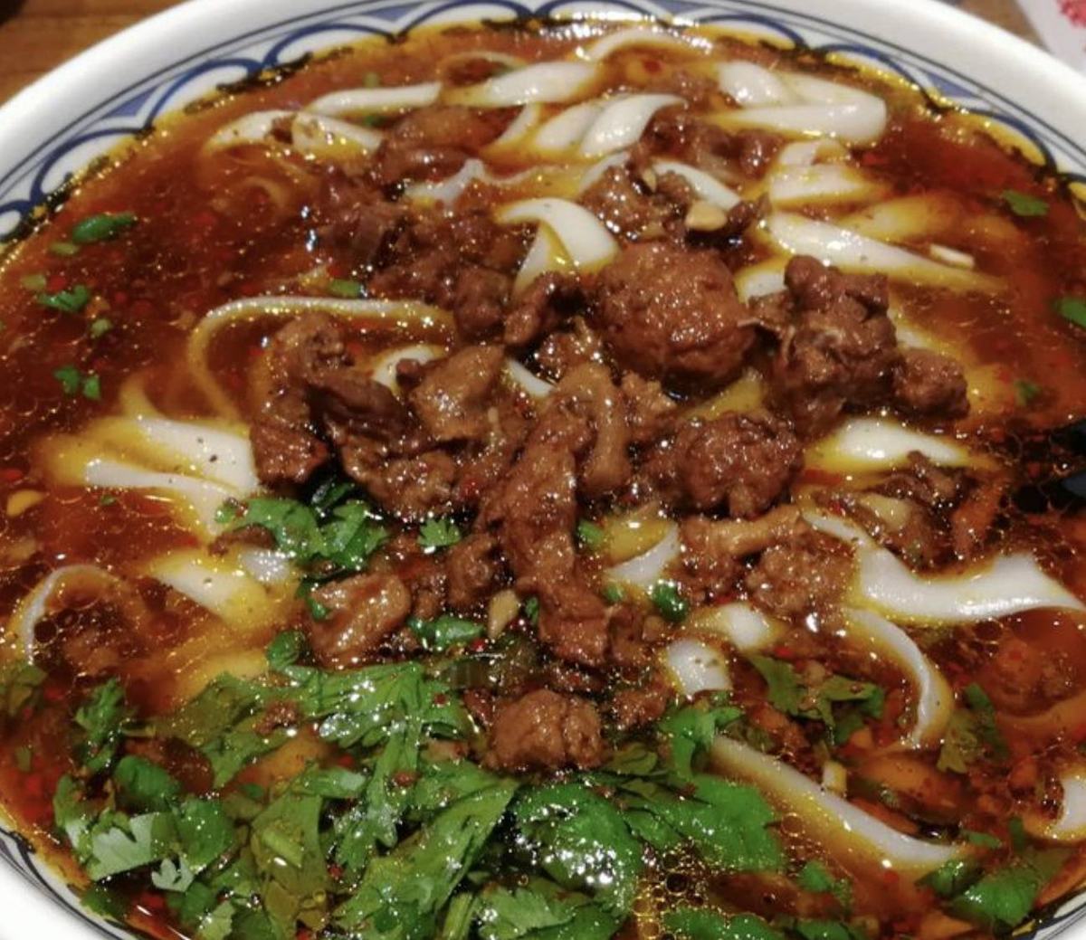 Knife-shaved noodle close up — broad, folded noodles thick through the middle and thin at the edges, sitting in glossy chilli-oil broth