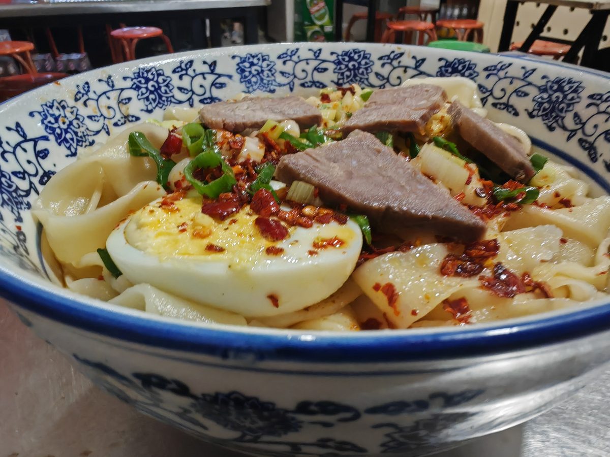 A bowl of knife-shaved noodles on the table of a small Chinese noodle shop, red plastic stools pushed back behind it
