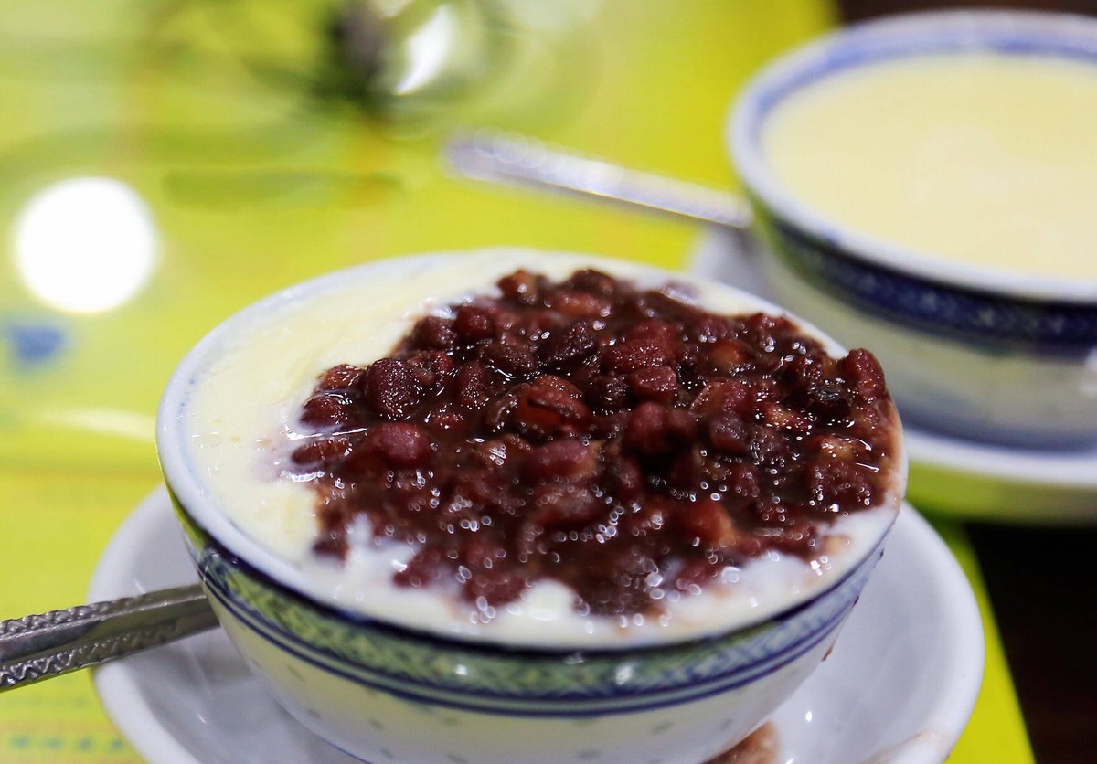 Two bowls of double-skin milk on a laminate table in a Cantonese dessert shop, one topped with red beans