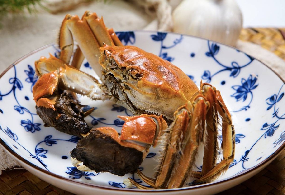 A mitten crab steamed the traditional way on blue-and-white porcelain, garlic bulb and bamboo mat behind it, served at the table as the starting point the cure transforms