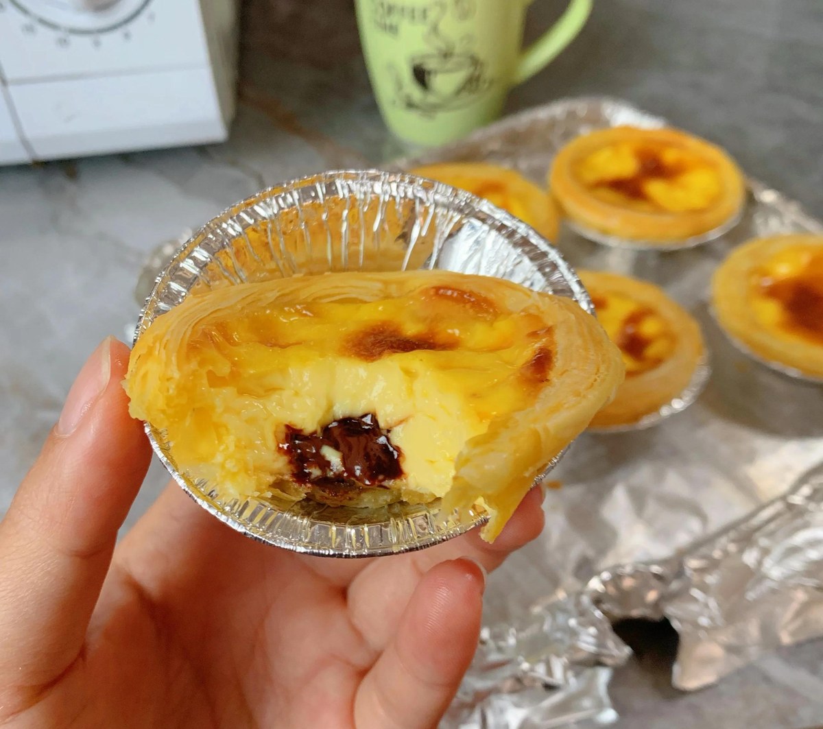 A hand holding an egg tart in its foil cup, a mug of milk tea within reach