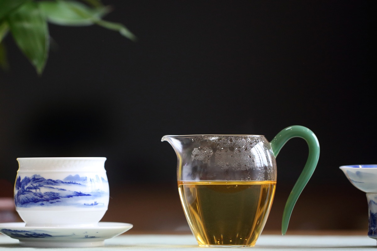 A glass fairness cup filled with amber tea on a wooden tea tray, a gaiwan resting behind it