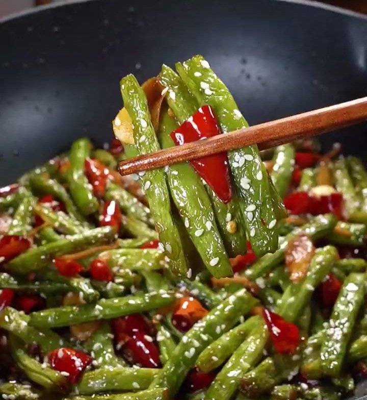 Gan Bian Si Ji Dou close-up — chopsticks lifting a single blistered bean, wrinkles and char marks visible, minced pork clinging to the surface, chili flecks scattered
