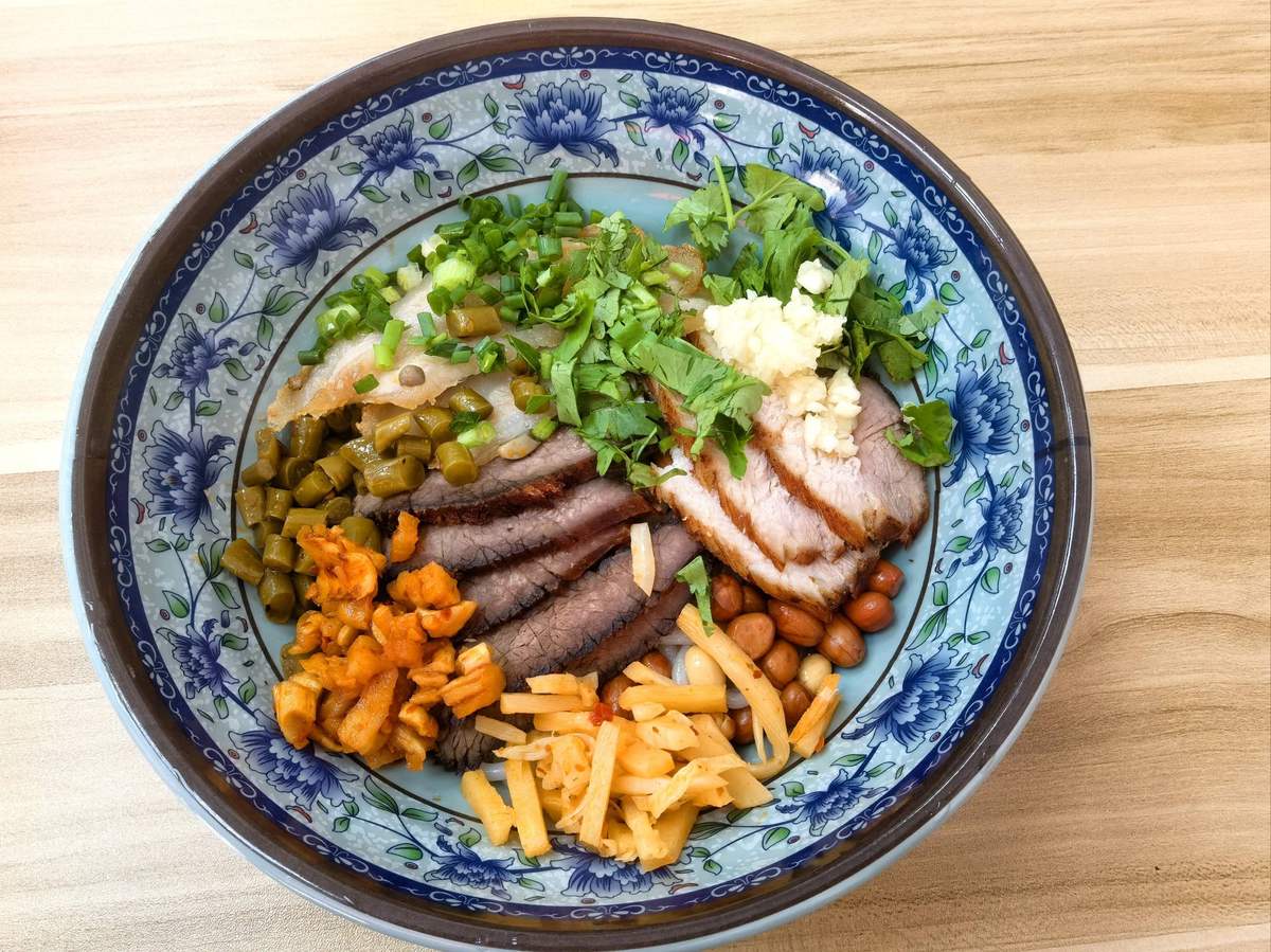 Guilin rice noodles served dry — flat white rice noodles under dark master braise, sliced braised beef and blistered 锅烧 pork belly, with pickled long beans, peanuts, cilantro and scallion in a blue-and-white bowl on a bare wooden table