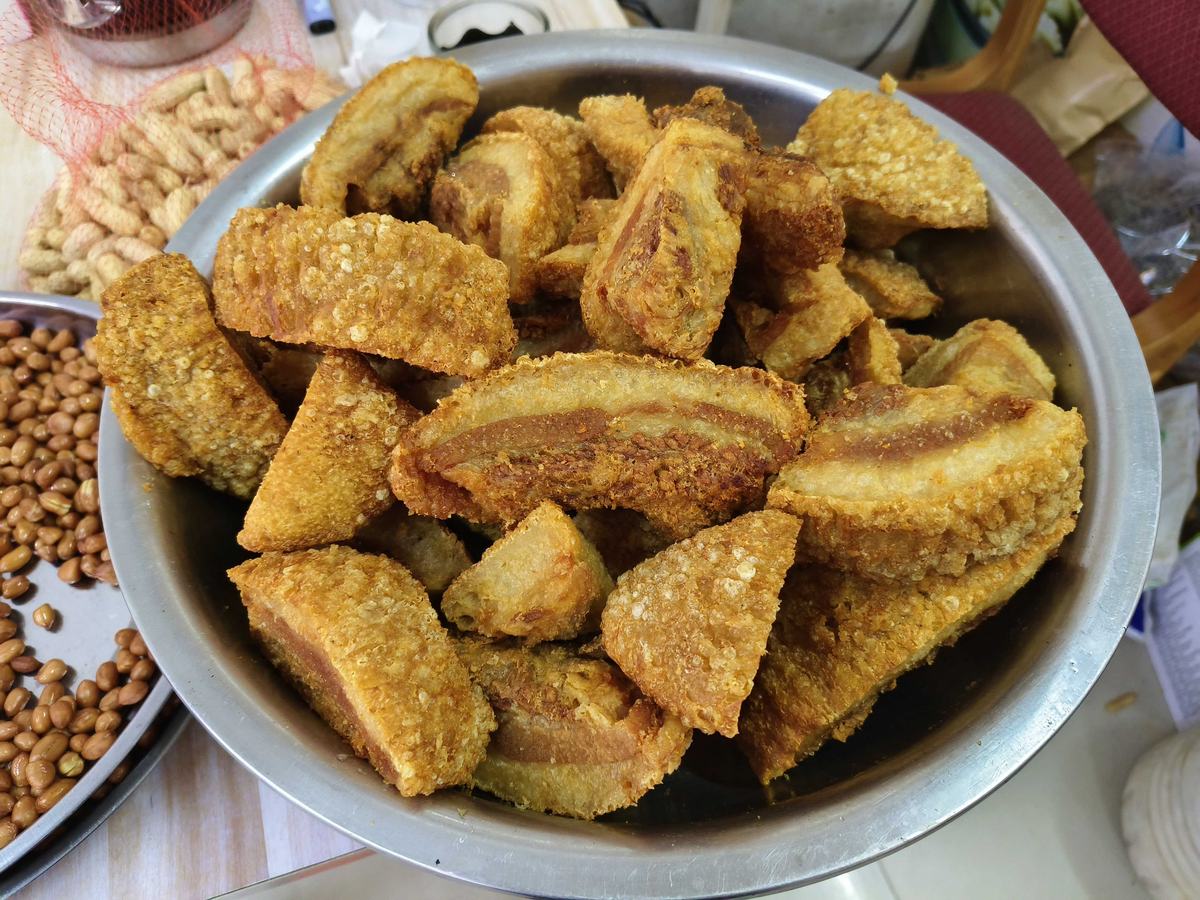 锅烧 close-up — slabs of twice-fried pork belly with puffed golden crackling skin and rendered fat, resting in a well-used steel bowl beside a plate of roasted peanuts