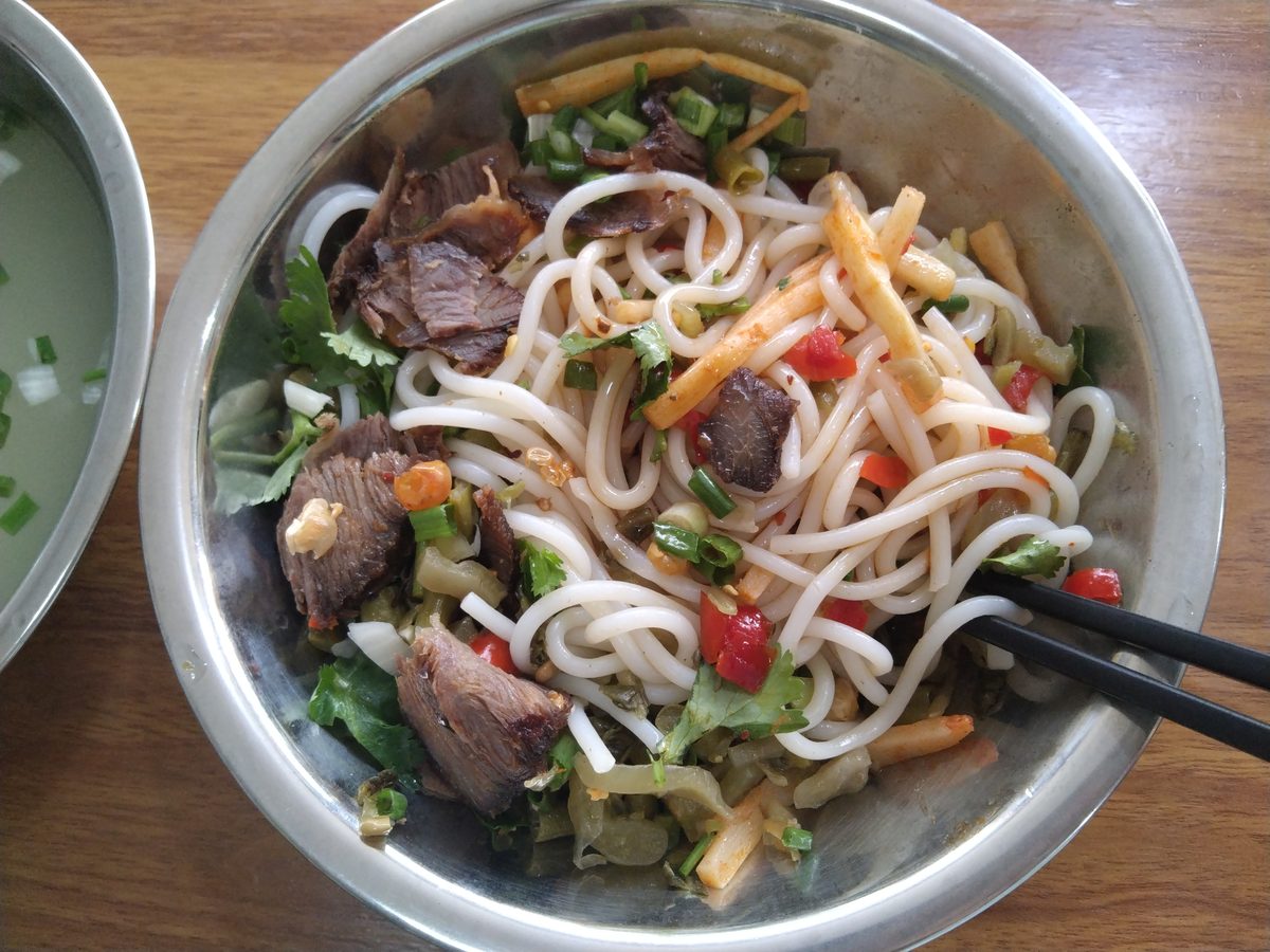 The second half of the bowl — steel bowl of dry-tossed Guilin rice noodles with braised beef and pickles, black chopsticks across the rim, and the small side bowl of clear pork-bone broth waiting to be poured in