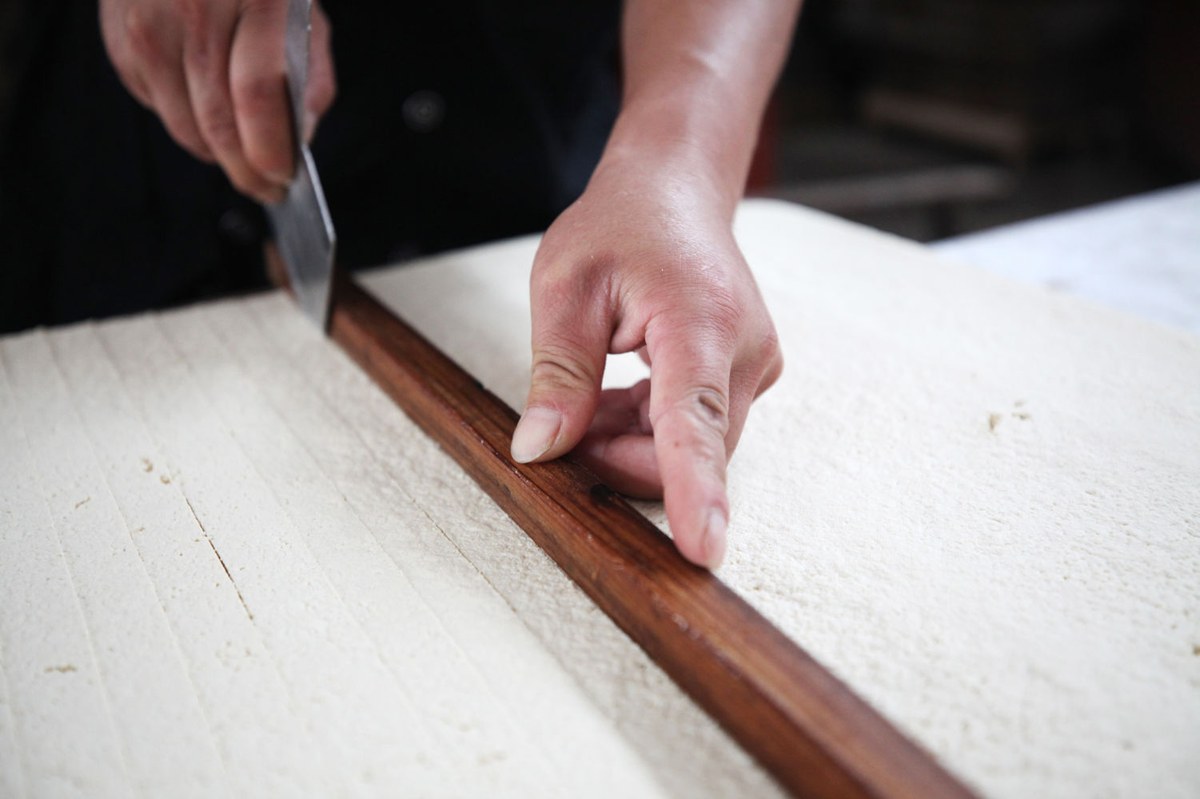 Hairy tofu being made — a maker portioning a tray of mold-covered tofu along a wooden straightedge in a Huizhou workshop