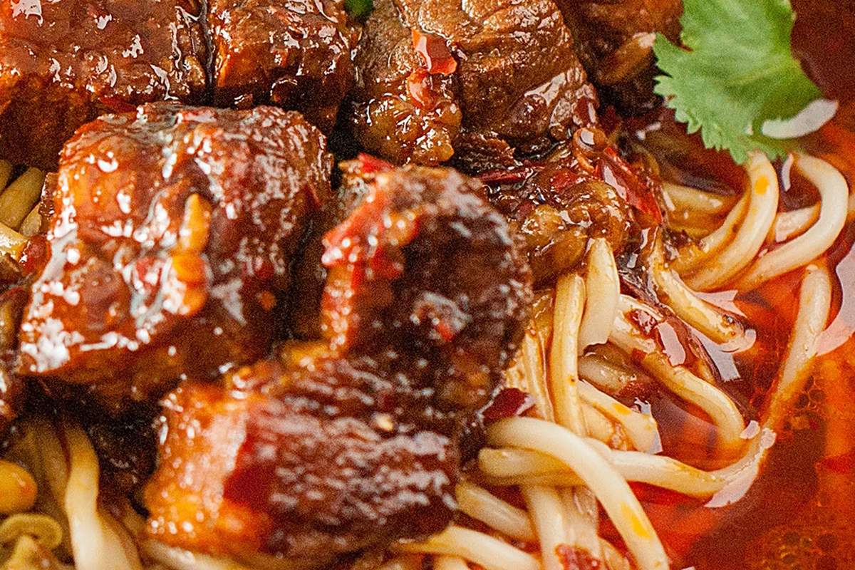 Hong Shao Niu Rou Mian close-up — glistening chunks of braised beef shank with tendon, dark broth, steam rising