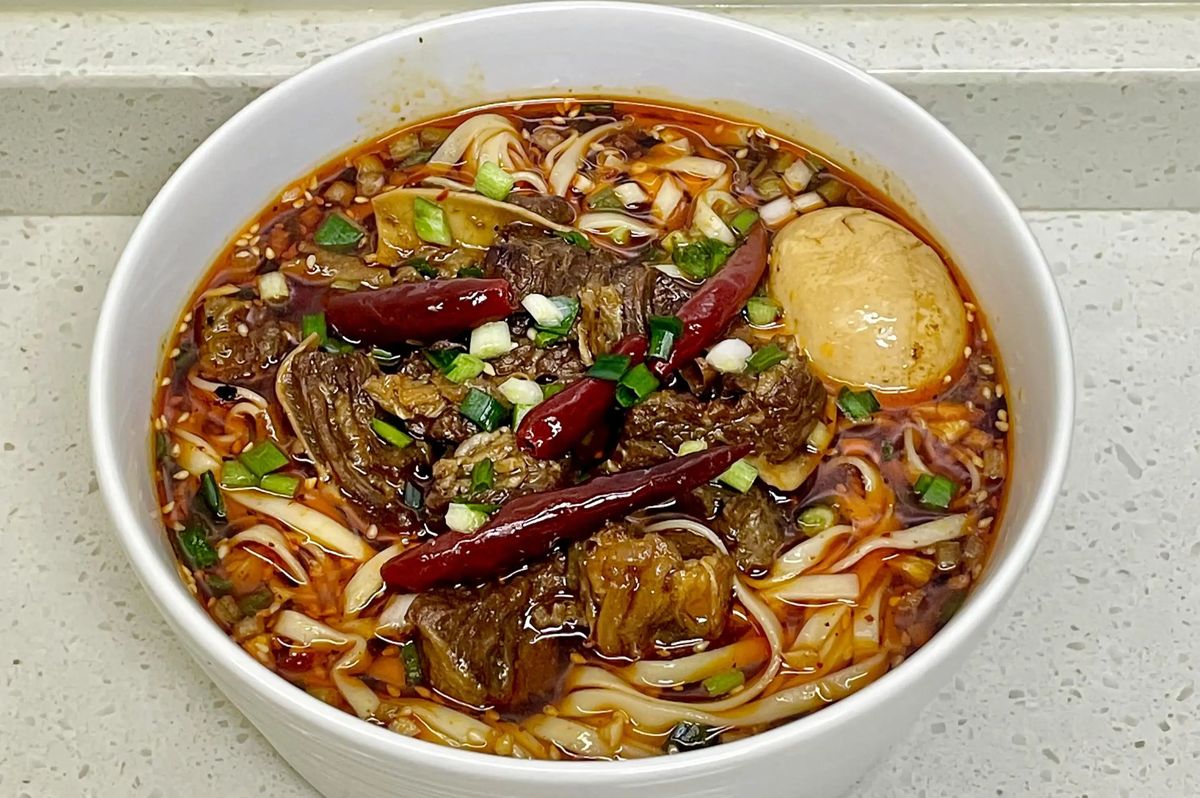 Hong Shao Niu Rou Mian served in a traditional bowl — thick noodles, beef chunks, dark broth, pickled mustard greens on the side