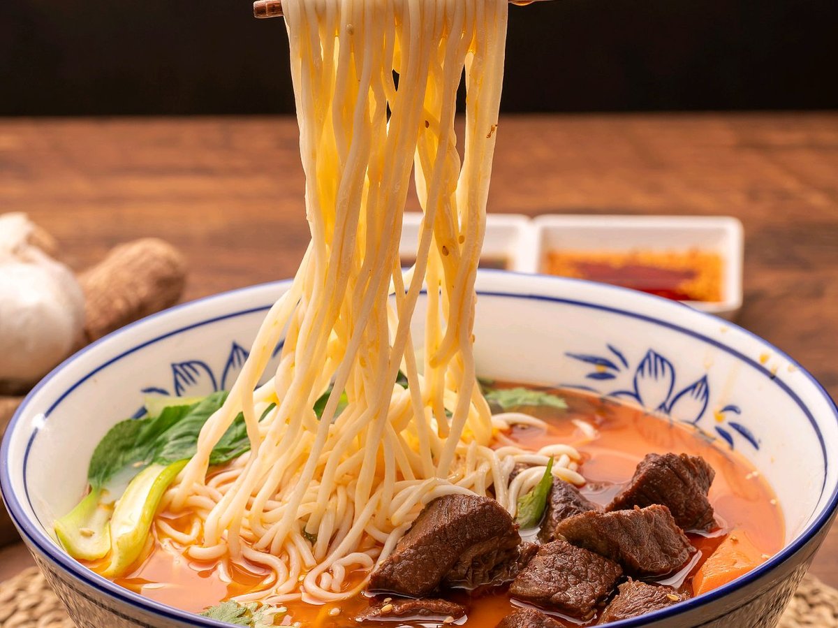 Hong Shao Niu Rou Mian being eaten — chopsticks lifting noodles from dark broth, steam visible, cozy shop setting