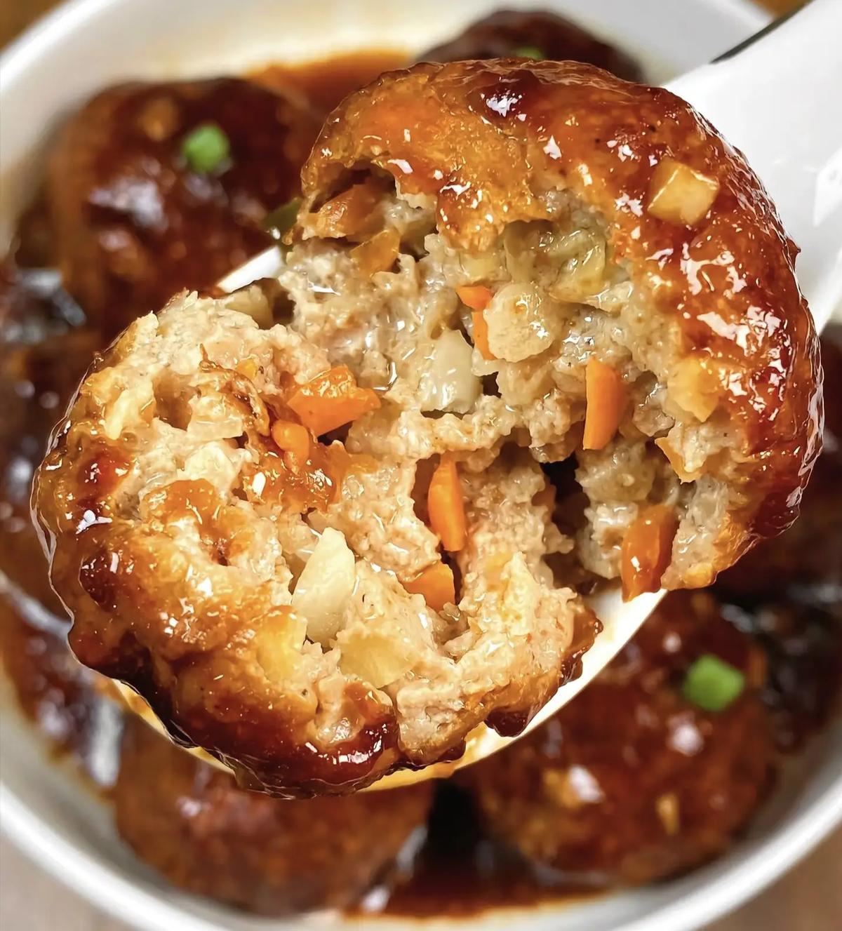 Close-up — spoon breaking open a lion&rsquo;s head meatball, glistening interior, water chestnut flecks visible, sauce pooling