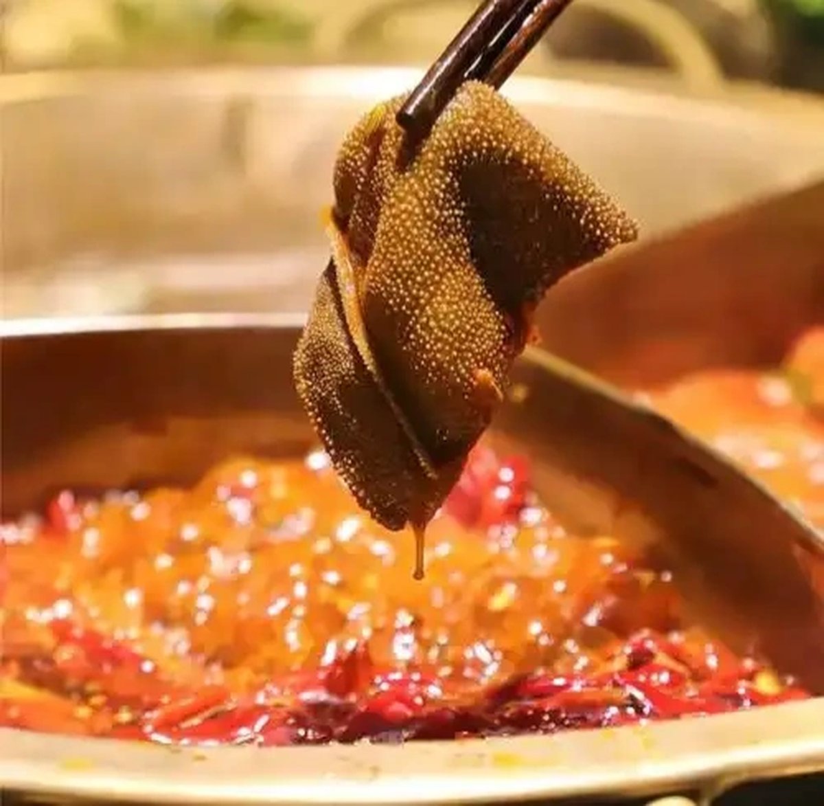 Hot pot close-up — chopsticks lowering a slice of marbled beef into bubbling chili oil, the meat turning from red to brown in seconds