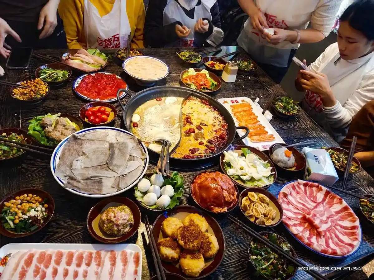 Hot pot table — six people around a steaming cauldron, chopsticks everywhere, faces flushed from spice, beer bottles on the table, the controlled chaos of a meal in progress