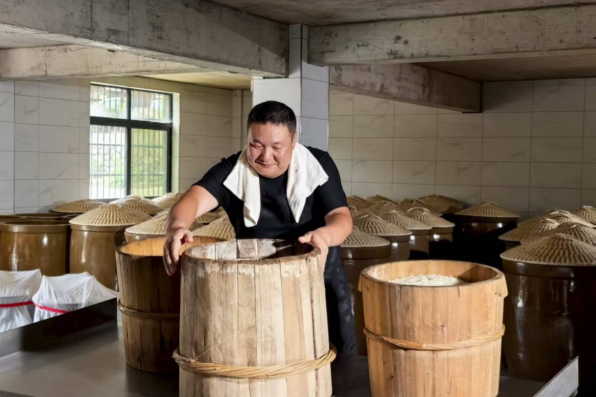 A worker lifting a wooden steamer of cooked glutinous rice, rows of fermentation jars behind him