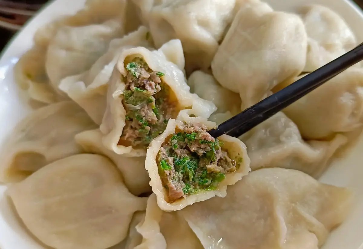 Jiaozi close-up — a single boiled dumpling held in chopsticks, translucent skin revealing the pork and cabbage filling inside, a drop of black vinegar on the surface