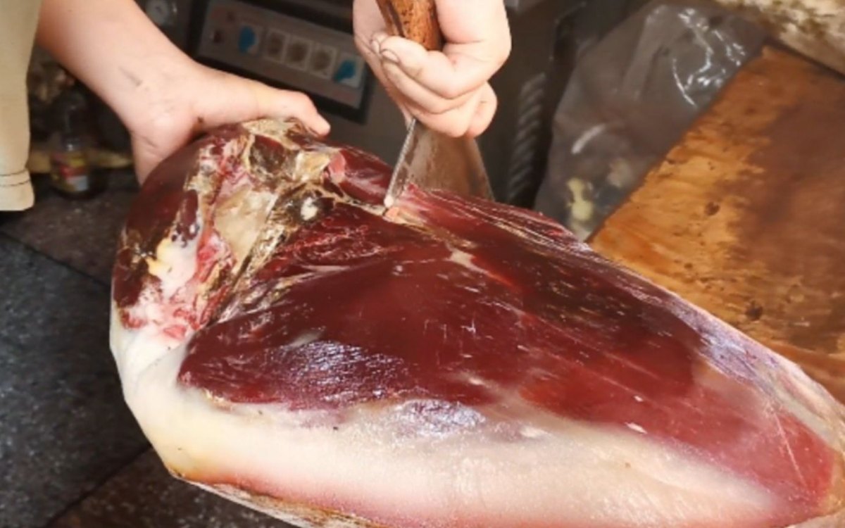 Two hands pushing a knife through a dense cured ham leg, the cut face deep red against a thick layer of white fat