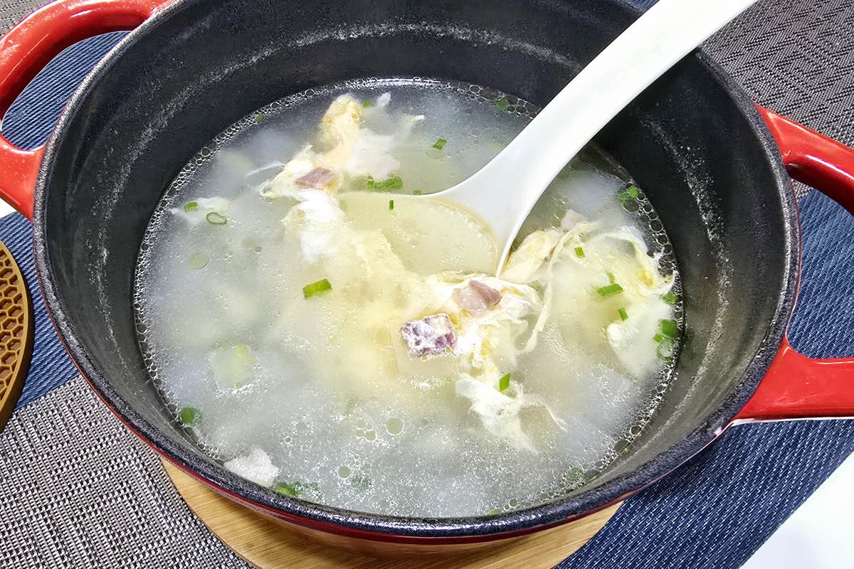A pot of winter melon soup with cubes of cured ham and pork ribs in clear broth, set on a woven mat
