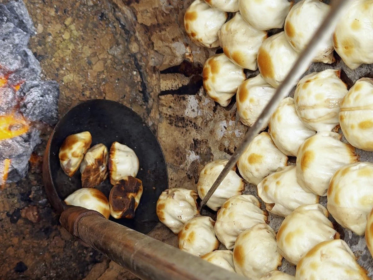 Inside a tandoor — pale buns stuck to the scorched clay wall, coals glowing below and an iron hook in the foreground