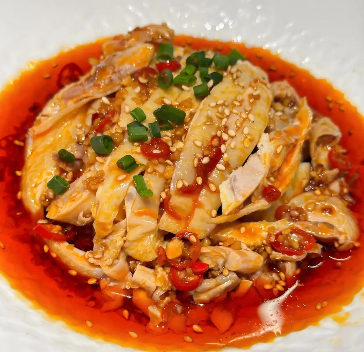 Kou Shui Ji close-up — a single cross-section slice of cold poached chicken with skin intact, glistening chili oil, sesame seeds catching the light