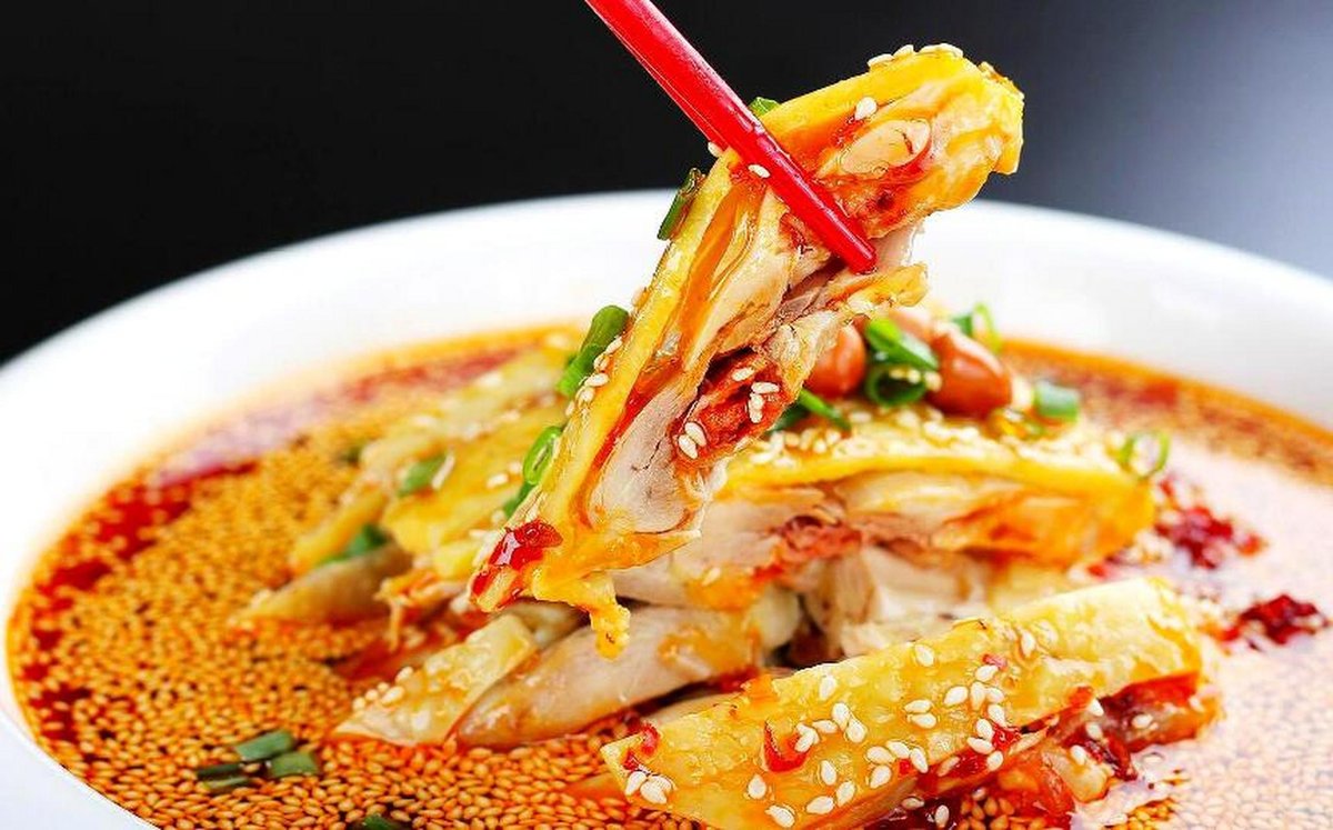 Kou Shui Ji — the finished dish on a white plate, chopsticks lifting a piece of chicken from the crimson chili oil pool, Sichuan restaurant table