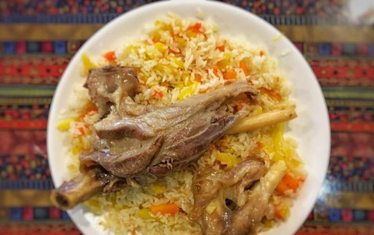 Lamb pilaf — a bone-in lamb shank resting on a mound of carrot-golden rice, white plate on a patterned Uyghur tablecloth