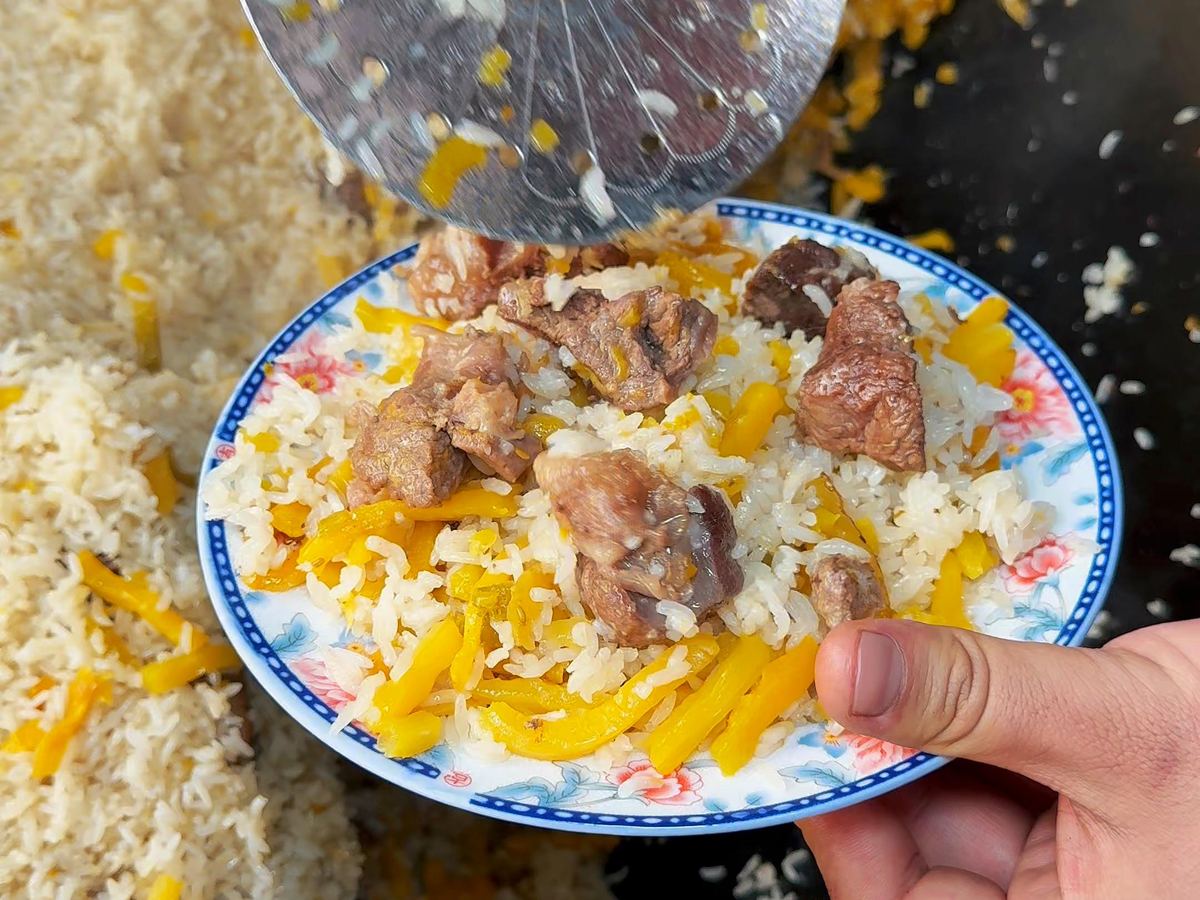 Lamb pilaf being served from a wide pot — a spoon lifting rice, carrot and lamb onto a plate at the edge of the frame
