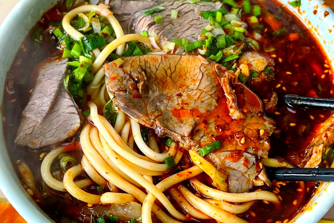 Lanzhou Beef Noodles close-up — hand-pulled noodles glistening with chili oil, sliced beef, and fresh herbs
