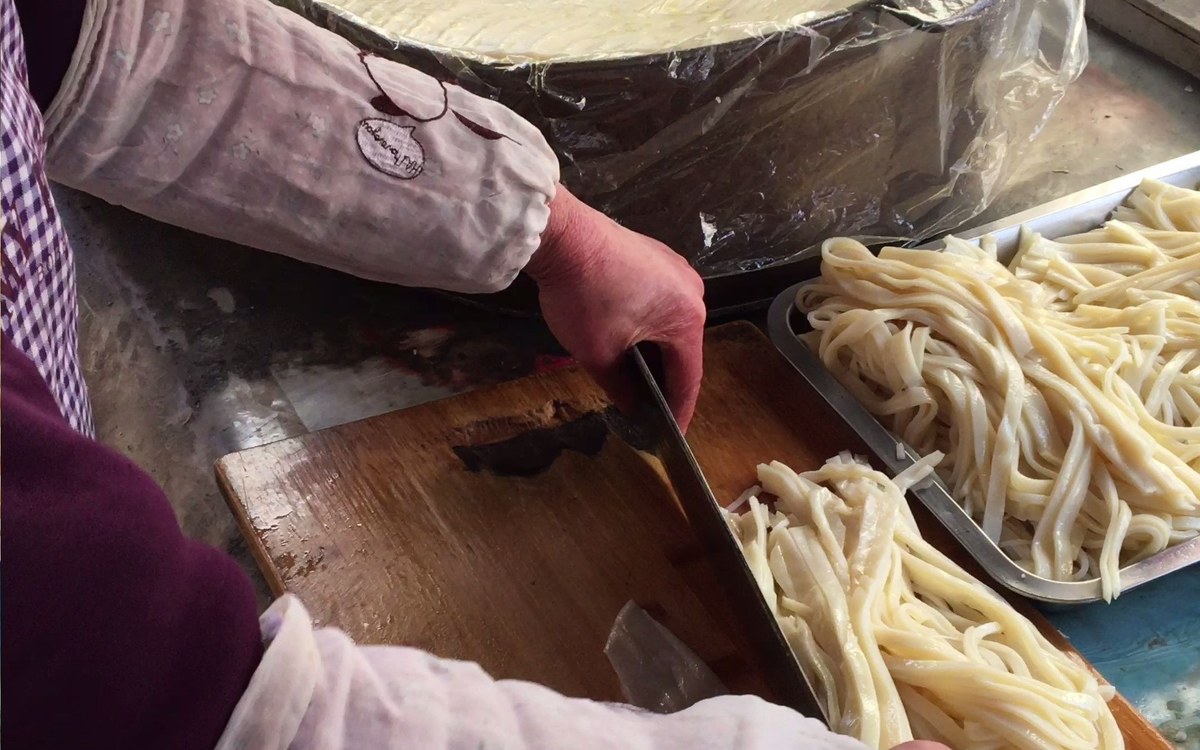 Liangpi being cut — a steamed starch sheet sliced into finger-width ribbons with a cleaver on a worn wooden board