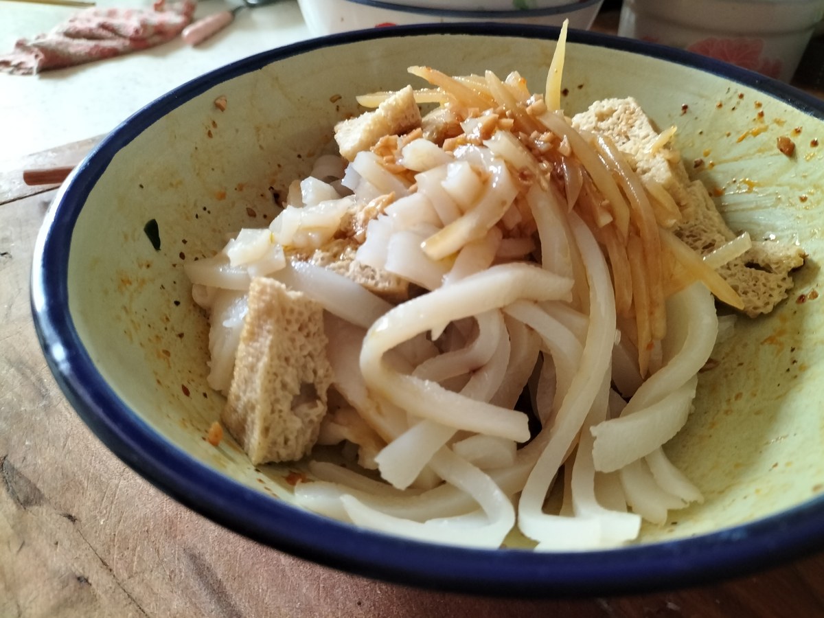 Close-up of liangpi with porous cubes of steamed wheat gluten, cucumber matchsticks and chile oil pooling in an enamel bowl