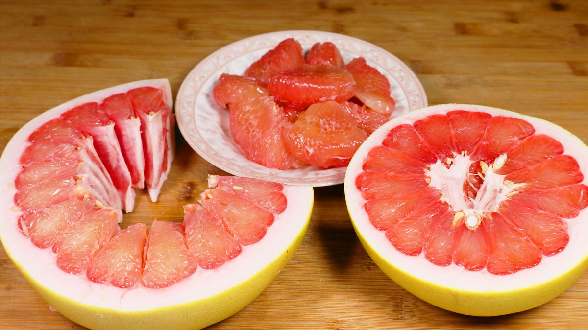 A halved pomelo on a wooden cutting board in a home kitchen, segments broken apart by hand beside a plate of peeled pulp