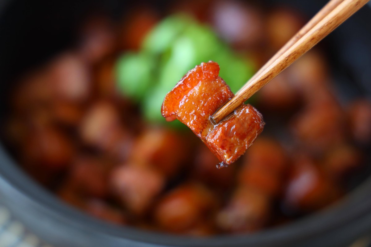 Red-braised pork close-up — one cube in chopsticks, the five layers of belly showing through the glaze