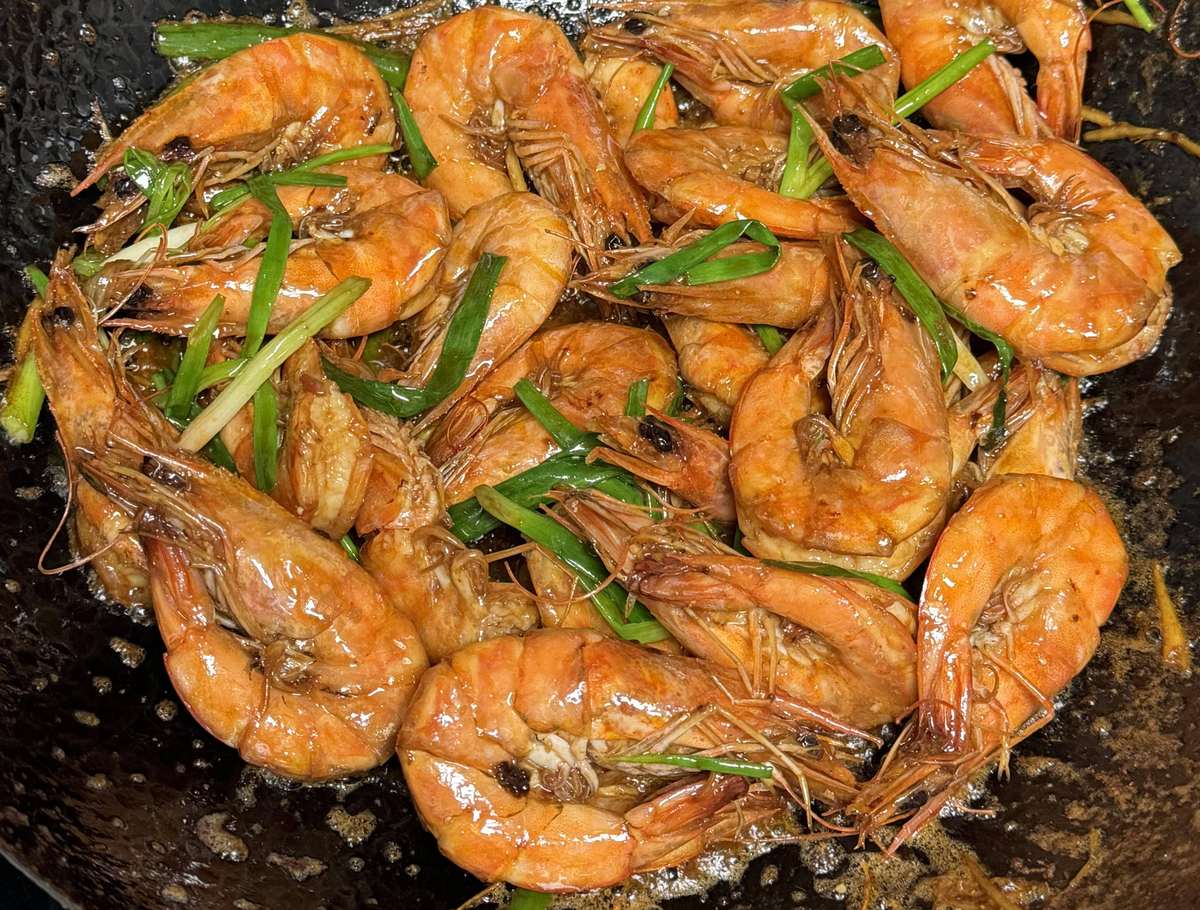 Whole prawns turning in a dark wok, heads and shells glossy with red oil, scallion scattered through