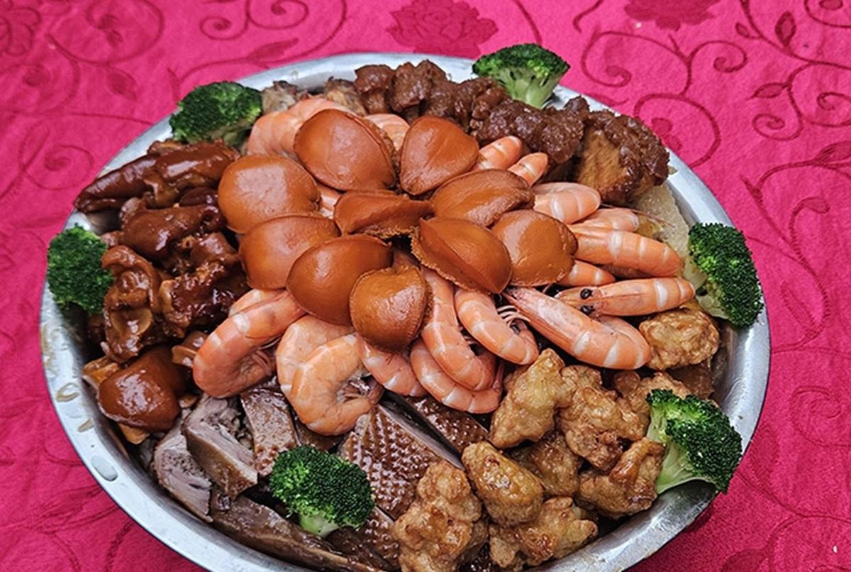 A whole poon choi (盆菜) — a stainless steel basin piled with abalone, prawns, braised pork, roast pork and broccoli, set down on a patterned tablecloth