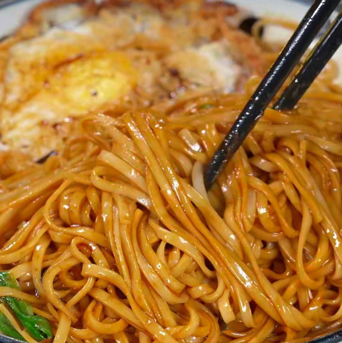 Close-up of scallion oil noodles — dark soy-coated strands glistening under kitchen light, flecks of fried scallion scattered across the surface