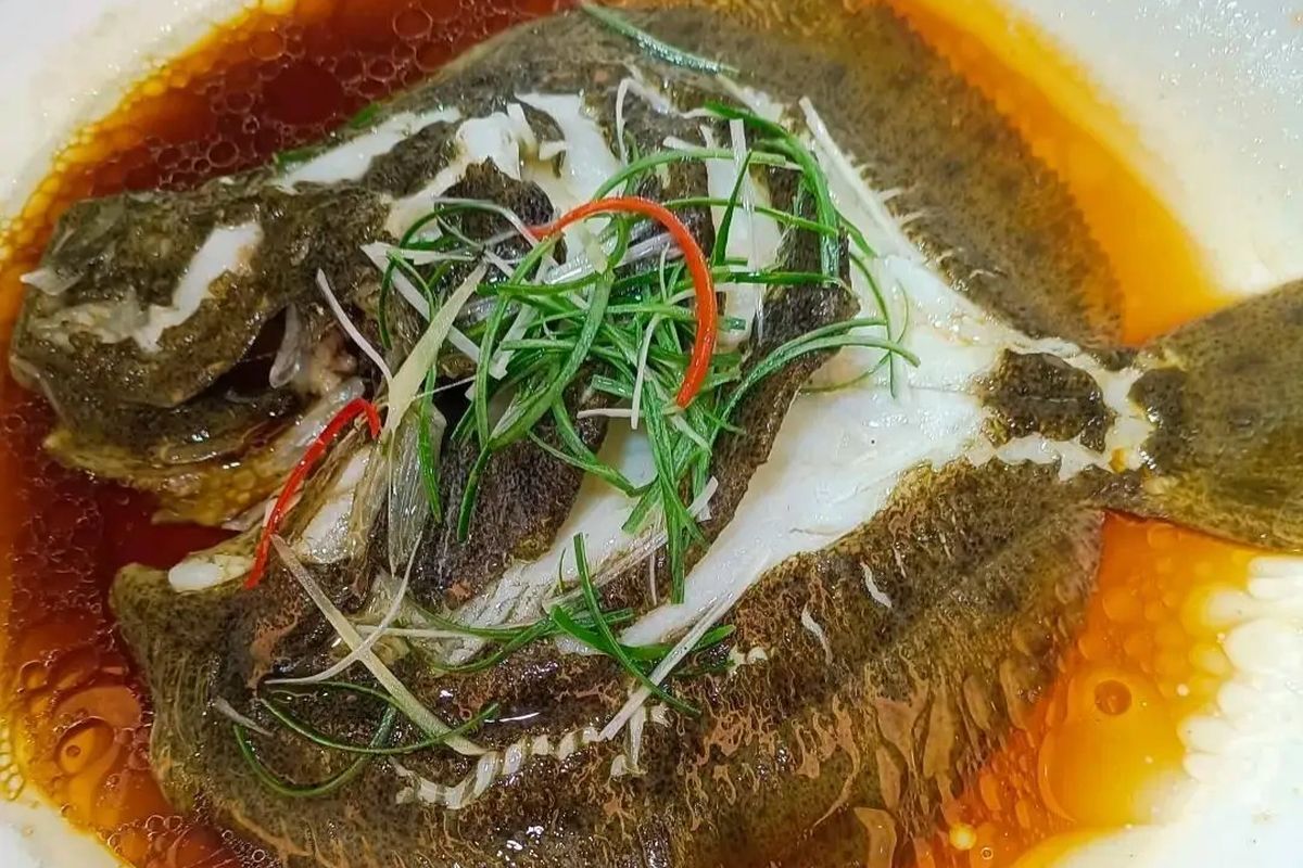 Steamed fish Cantonese style — whole sea bass on an oval platter, ginger threads and scallion ribbons draped across the body, soy sauce pooling beneath, chopsticks hovering